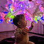Aurora joined the competition — help win amazing prizes! baby, child, christmas_tree, ornament, lights, colorful, indoor, holiday, decorations, white_tree, curious, infant, soft_blanket, festive, celebration, glowing, sitting, holding, reaching, profile