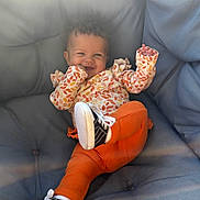 Aurora is registered to the contest to win money with this photo: baby, child, smiling, curly_hair, orange_pants, sneakers, couch, cushion, indoor, happy, playful, infant, clothing, portrait, person, cute, relaxing, toddler, seat, cozy