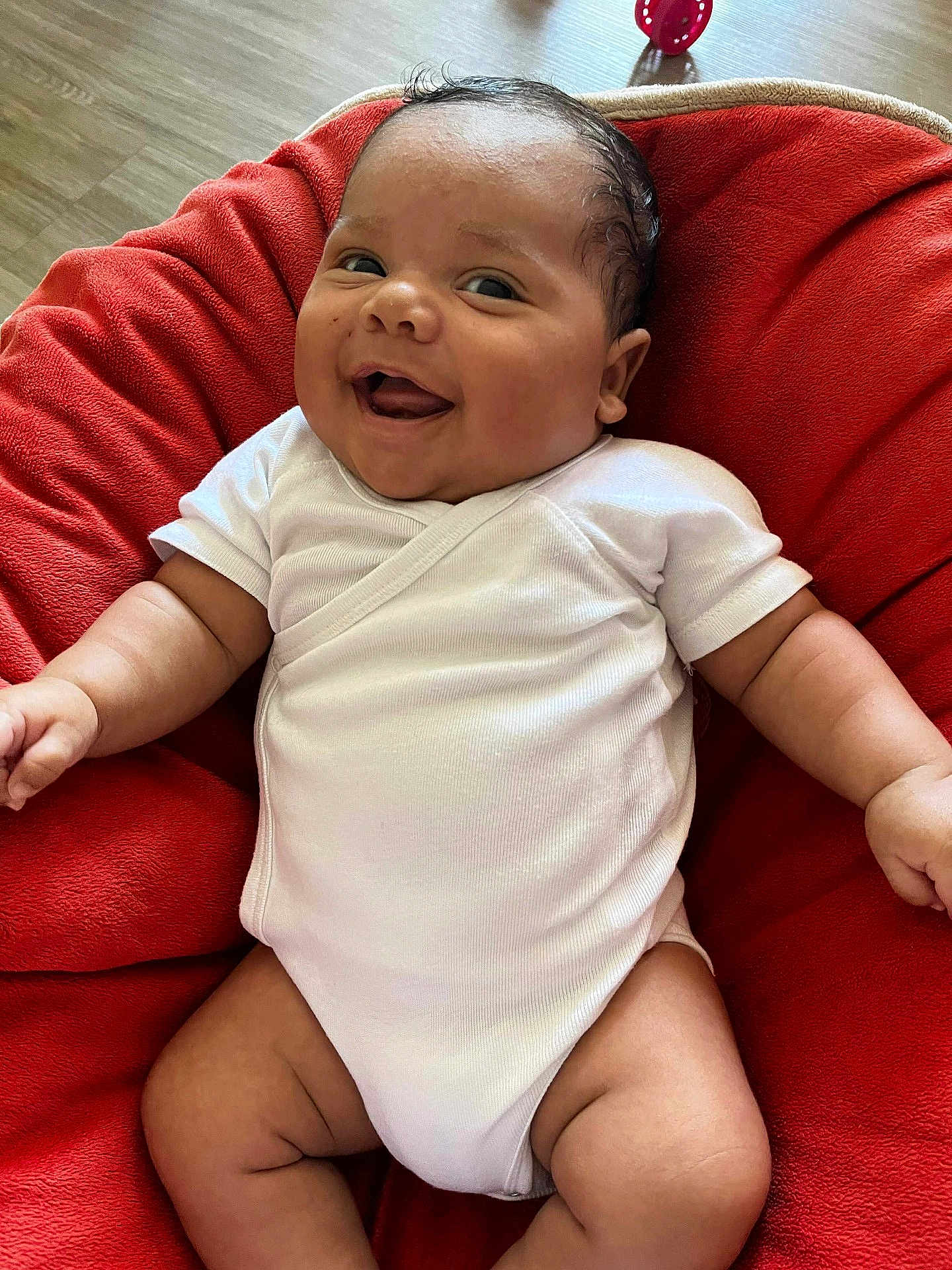 Louan participe au concours pour gagner de l'argent avec cette photo : baby, infant, smile, white_onesie, red_cushion, happy, chubby_arms, chubby_legs, indoors, floor, wooden_floor, cute, portrait, child, human, skin, person, sitting, soft_fabric, cozy
