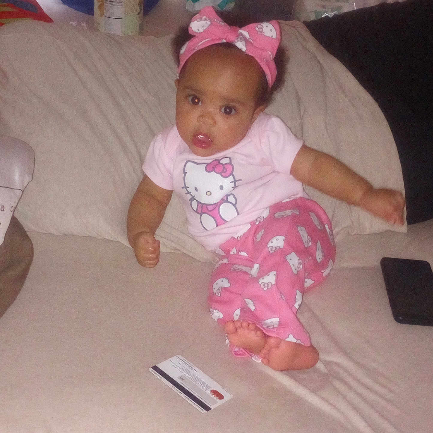 La'Skia joined the competition — help win amazing prizes! baby, casual, child, couch, credit_card, curious, face, footwear_none, headband, hello_kitty, home, indoor, infant, looking_at_camera, pajamas, person, pink_clothing, sitting, smartphone, soft_furniture