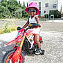 child, toddler, toy_motorbike, red_bike, pink_hat, sunglasses, outdoor, sunny, asphalt, training_wheels, smiling, summer_clothing, shorts, tank_top, shoes, urban, greenery, building, daytime, play