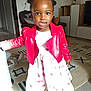 child, girl, red_jacket, floral_dress, smiling, indoors, living_room, carpet, furniture, television, cushion, hair_buns, playful, person, home, cute, portrait, standing, young, happy