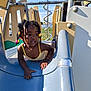 child, girl, playground, slide, outdoor, daytime, smiling, hair_beads, yellow_dress, plastic_slide, metal_structure, fence, grass, blue_sky, recreation, fun, active, person, kids_playing, summer