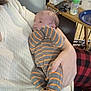 Theodore is registered to the contest to win money with this photo: baby, infant, striped_clothing, onesie, adult_arm, holding, indoor, table, baby_bottle, clothing, person, face, hand, plaid_pants, furniture, bottle, feeding, resting, newborn, casual