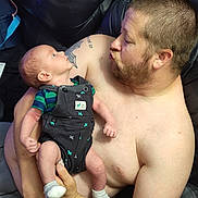 Theodore is registered to the contest to win money with this photo: baby, man, shirtless, beard, tattoo, overalls, socks, couch, leather, holding, playful, interaction, infant, person, indoor, closeup, family, looking, expression, casual