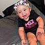 baby, infant, smiling, headband, onesie, pink, black_clothing, cheerful, happy, cute, bed, gray_bedspread, skin, hand, foot, person, indoor, child, young, sitting