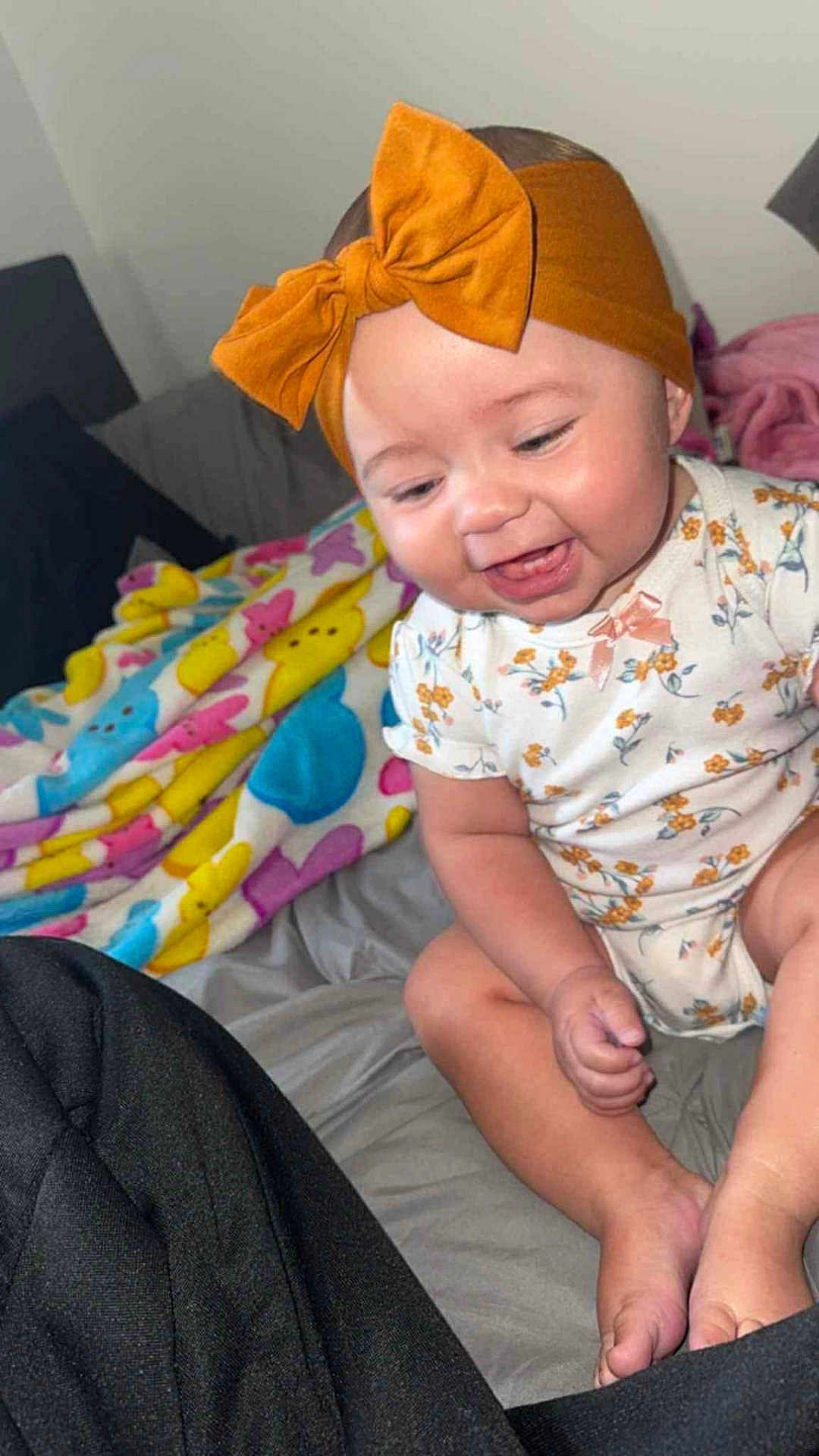 Londyn is registered to the contest to win money with this photo: baby, child, smiling, headband, bow, onesie, floral_pattern, blanket, colorful, bed, indoors, happy, cute, infant, sitting, person, skin, foot, hand, face