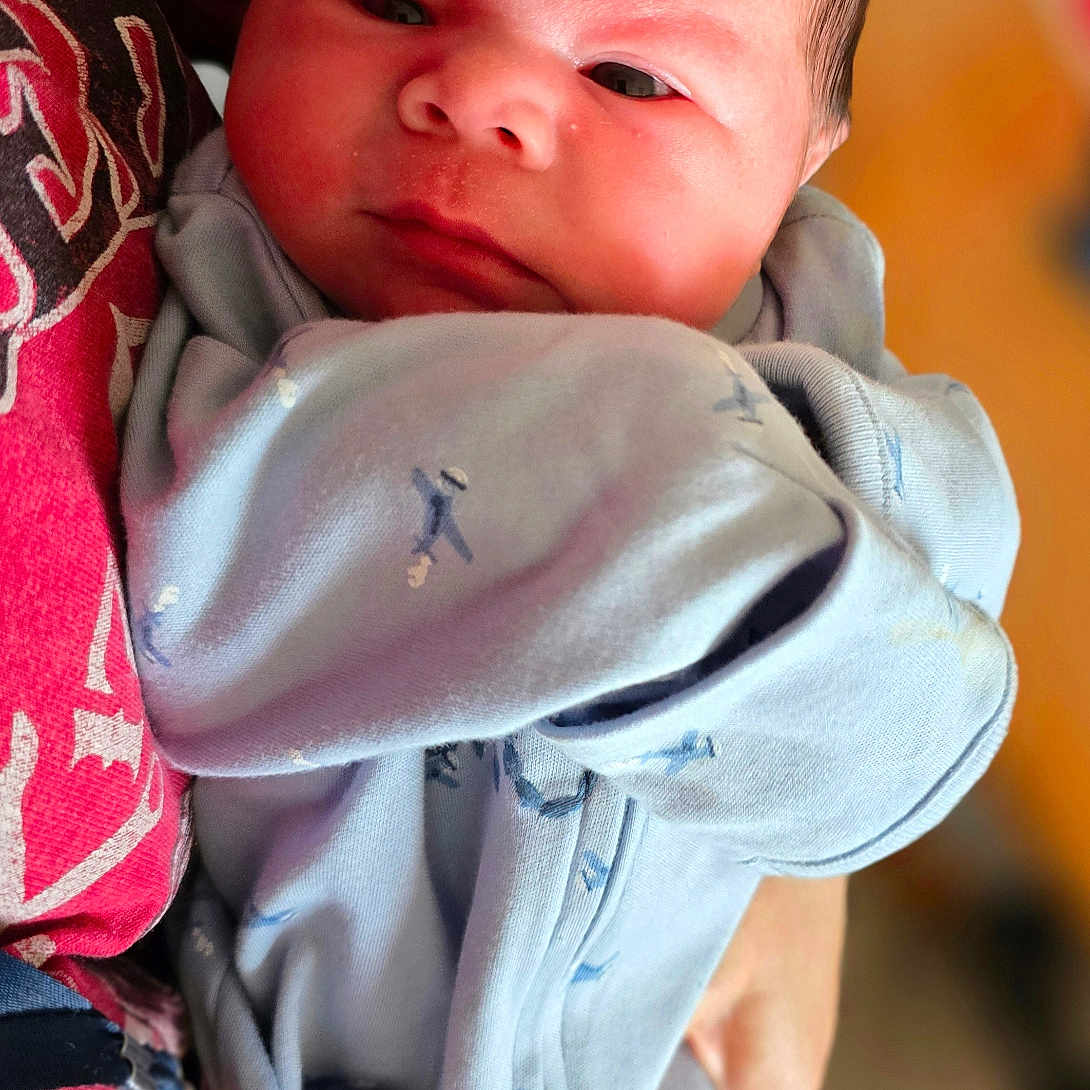 Zaylen is registered to the contest to win money with this photo: baby, newborn, infant, onesie, clothing, person, holding, red_shirt, closeup, face, portrait, indoor, human, cute, child, newborn_baby, baby_clothes, baby_face, soft_lighting, warm