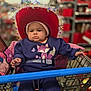 baby, shopping_cart, hat, purple_clothing, fox_illustration, tinsel, store, blurred_background, child, serious_expression, pink_hat, belt, hand, toy, retail, indoor, person, clothing, seat, infant