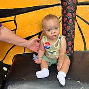 Noah is registered to the contest to win money with this photo: baby, child, pacifier, socks, tank_top, shorts, hand, bench, butterfly_wing_painting, face, blue_eyes, skin, camo_pattern, sitting, supporting_hand, voting_sticker, outdoor, expression, person, furniture