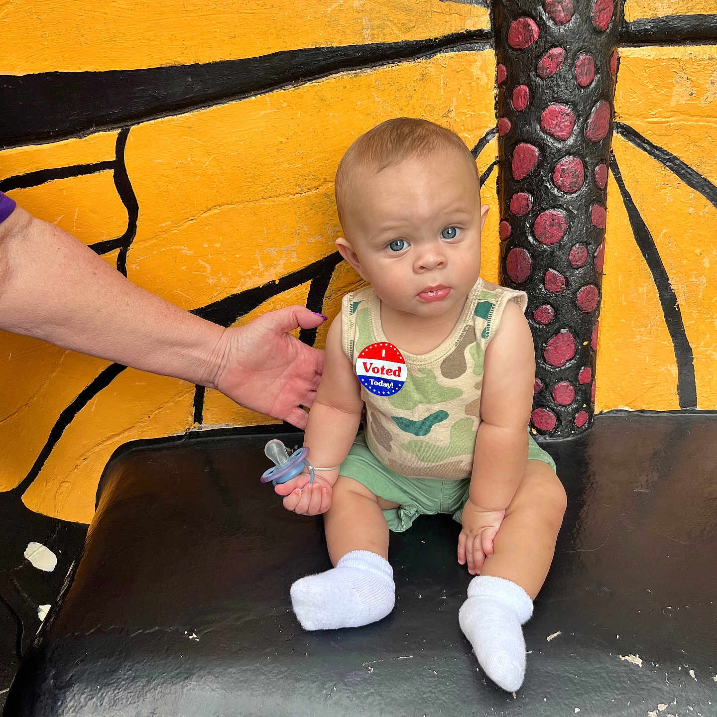 Noah is registered to the contest to win money with this photo: baby, bench, blue_eyes, butterfly_wing_painting, camo_pattern, child, expression, face, furniture, hand, outdoor, pacifier, person, shorts, sitting, skin, socks, supporting_hand, tank_top, voting_sticker