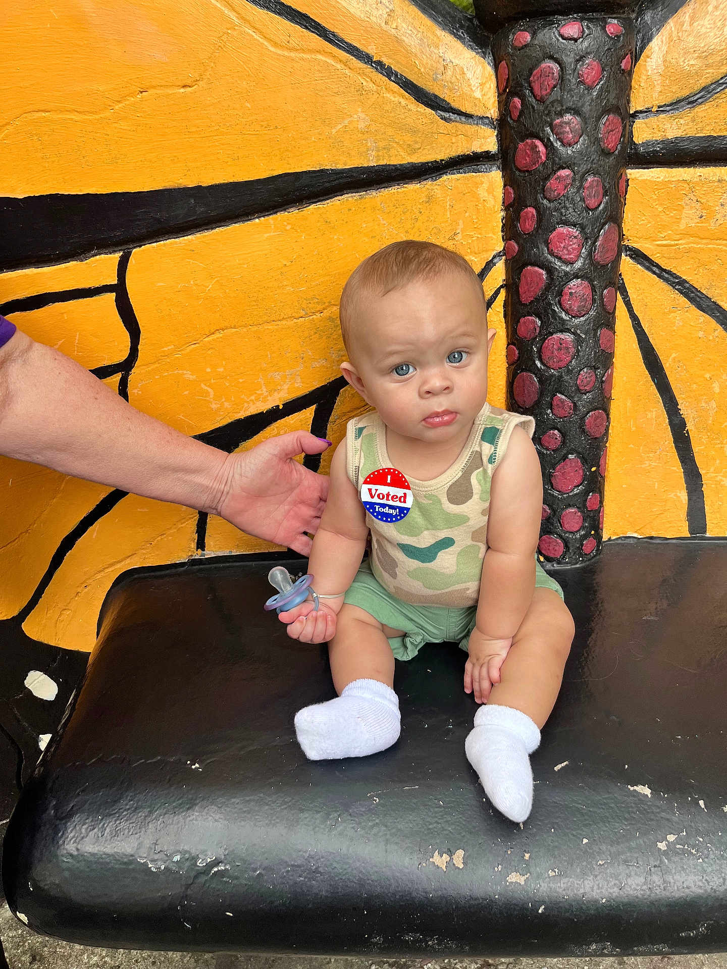 Noah is registered to the contest to win money with this photo: baby, child, pacifier, socks, tank_top, shorts, hand, bench, butterfly_wing_painting, face, blue_eyes, skin, camo_pattern, sitting, supporting_hand, voting_sticker, outdoor, expression, person, furniture