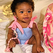 Ariyah is registered to the contest to win money with this photo: baby, infant, child, smiling, cute, curly_hair, pink_bow, blue_outfit, pacifier, beads, sitting, blanket, bedroom, soft_lighting, indoor, person, closeup, happy, accessory, bag