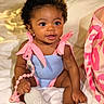 baby, infant, child, smiling, cute, curly_hair, pink_bow, blue_outfit, pacifier, beads, sitting, blanket, bedroom, soft_lighting, indoor, person, closeup, happy, accessory, bag