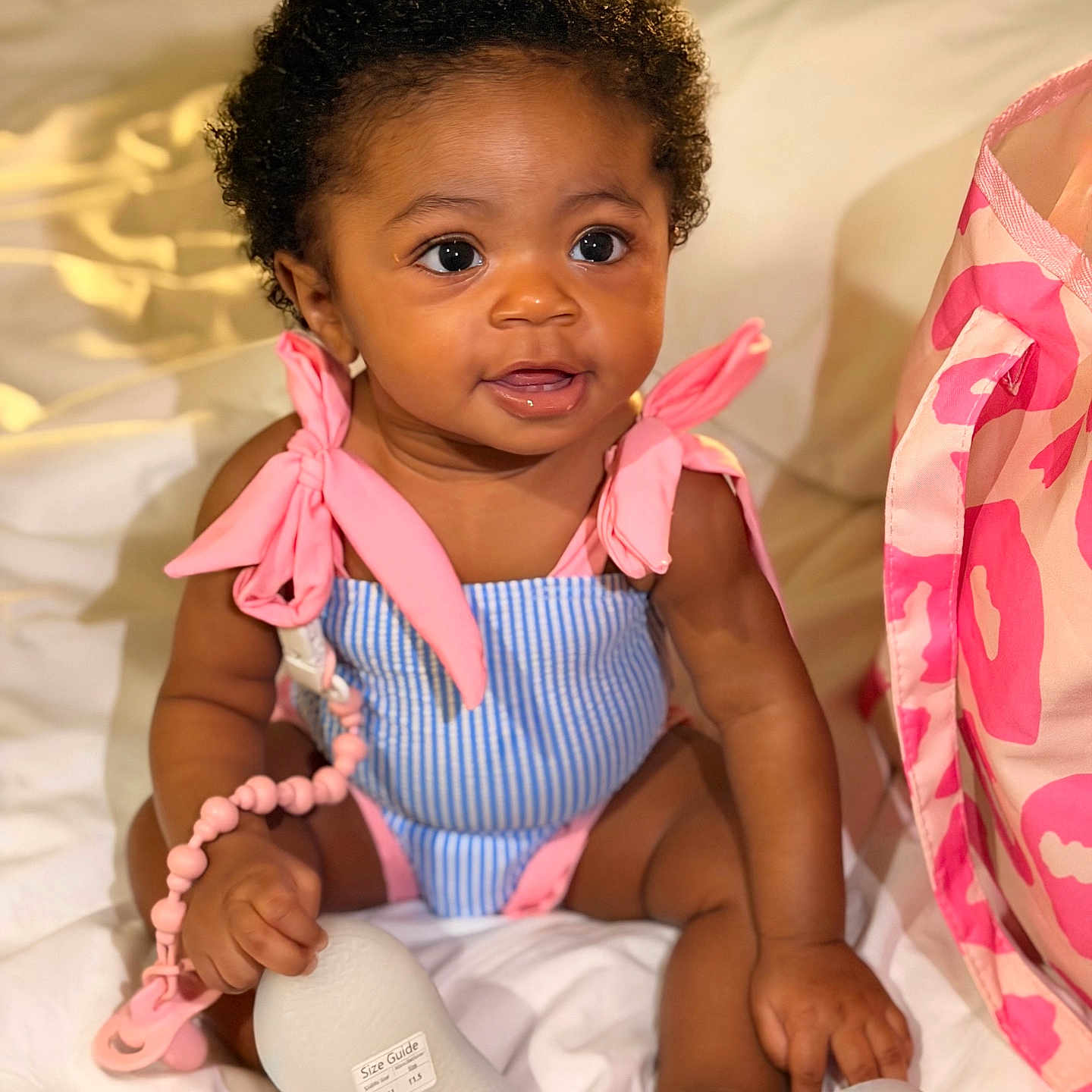 Ariyah is registered to the contest to win money with this photo: baby, infant, child, smiling, cute, curly_hair, pink_bow, blue_outfit, pacifier, beads, sitting, blanket, bedroom, soft_lighting, indoor, person, closeup, happy, accessory, bag