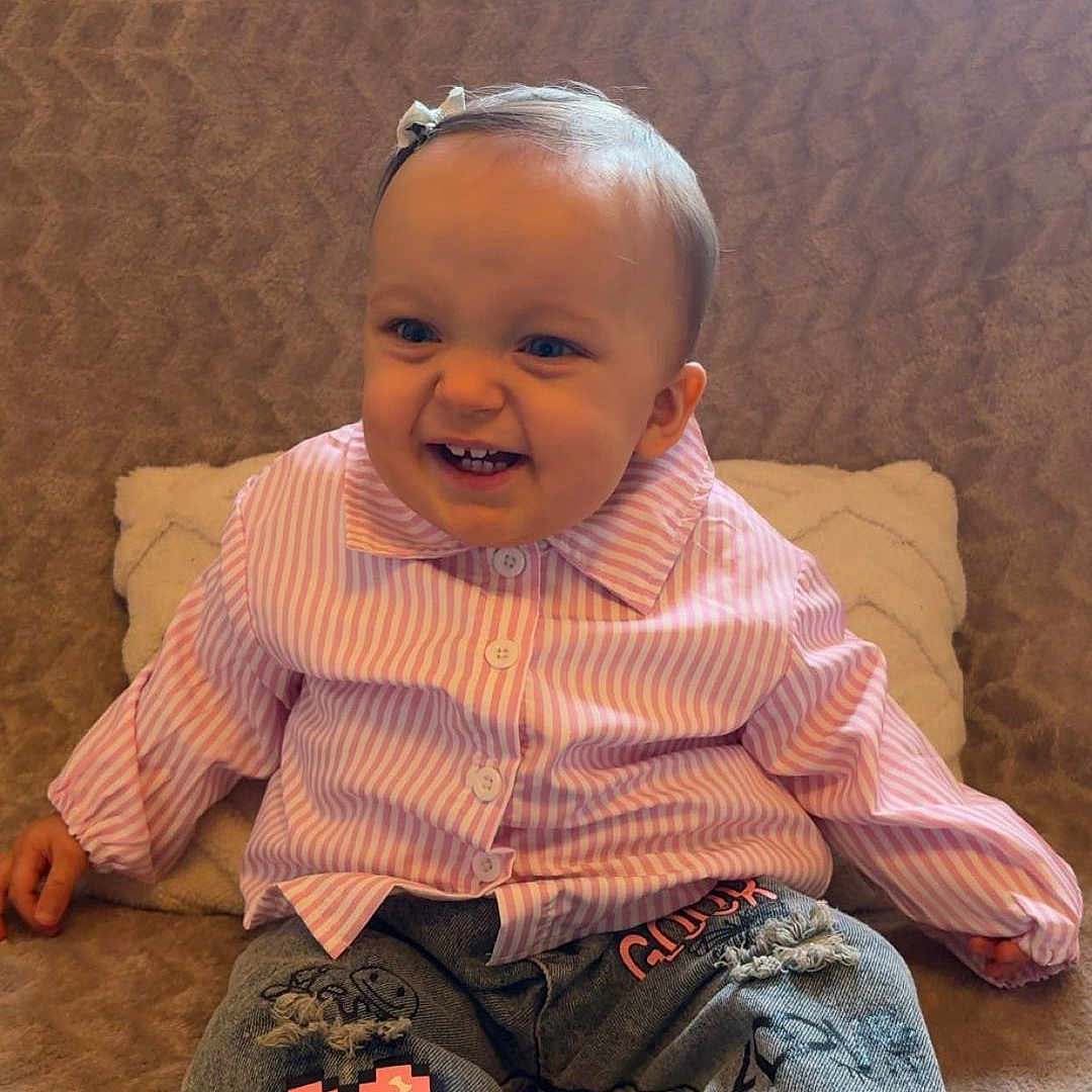 Ilyena participe au concours pour gagner de l'argent avec cette photo : baby, black_white_sneakers, casual_clothing, child, couch, cute, hair_bow, happy, indoor, jeans, pillow, pink_shirt, pixel_heart_patch, playful, portrait, sitting, smiling, striped_shirt, texture, toddler