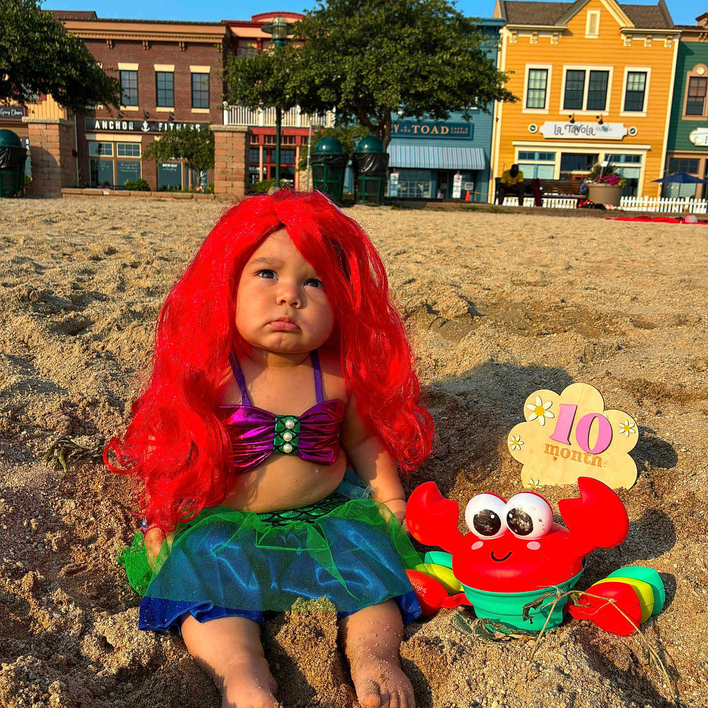 Ailany is registered to the contest to win money with this photo: child, baby, mermaid_costume, red_wig, crab_toy, sand, beach, boardwalk, outdoor, portrait, pouting, sitting, costume, colorful, sunlight, toy, birthday, ten_months, buildings, seaside