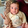 baby, child, blue_eyes, stuffed_animal, bunny, soft_toy, cream_shirt, animal_illustration, blanket, maroon_throw, indoor, portrait, cute, cozy, curious, hand, face, sitting, young, infant
