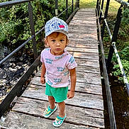 Marc a rejoint le concours — aidez-le/la à gagner de superbes lots ! child, toddler, bridge, wooden_bridge, cap, shorts, shirt, shoes, outdoor, nature, trees, greenery, water, stream, sunlight, curious, standing, person, summer, casual_clothing