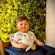Mateo a rejoint le concours — aidez-le/la à gagner de superbes lots ! toddler, child, high_chair, smile, baby, outdoor, greenery, leafy_background, sunlight, clothing, seatbelt, wooden_chair, face, happy, cute, portrait, summer, casual_wear, baby_toy, playful