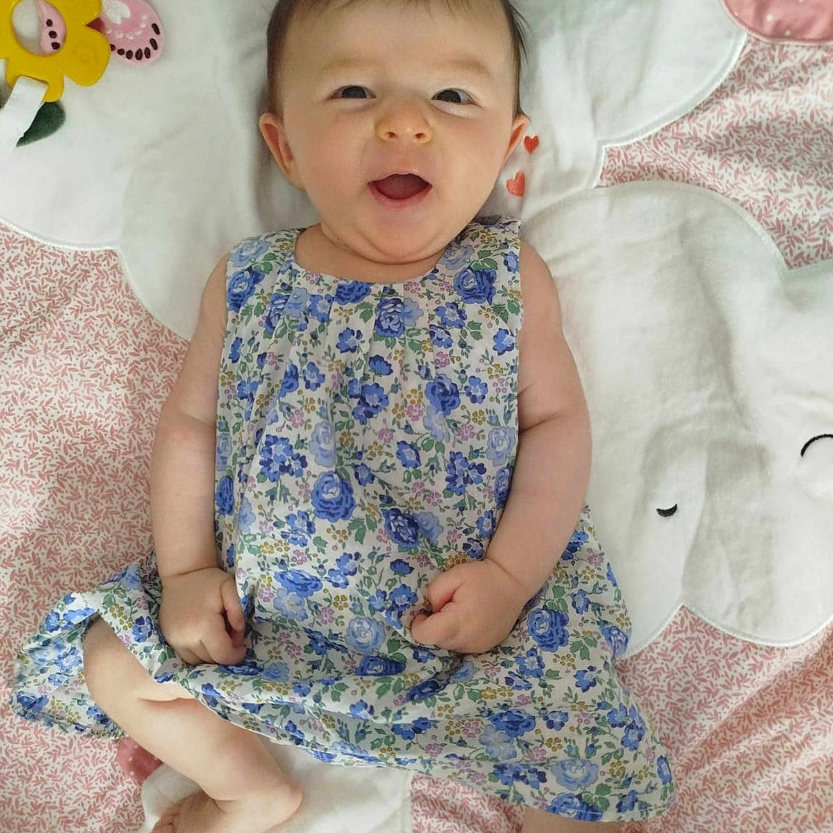 Mélody participe au concours pour gagner de l'argent avec cette photo : baby, blanket, child, cozy, cute, expression, face, feet, floral_dress, happy, indoors, infant, lying_down, patterned, portrait, skin, smiling, soft, toy, yellow_flower