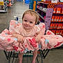 baby, child, shopping_cart, store, aisle, clearance_sign, pink_clothing, padded_cover, barefoot, smiling, blue_eyes, short_hair, retail, floor, shelves, products, orange_packaging, purple_display, baby_in_cart, indoor