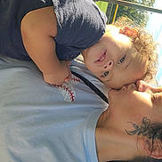 Adonis is registered to the contest to win money with this photo: child, adult, outdoor, sunlight, grass, fence, affection, kiss, tattoo, casual_clothing, portrait, person, family, love, happy, closeup, nature, daylight, curly_hair, smile