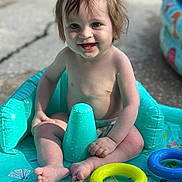Laurence joined the competition — help win amazing prizes! toddler, child, inflatable_pool, water_toy, smiling, sitting, summer, outdoor, play, fun, barefoot, blue, yellow, toy_ring, happy, young_child, water, splash, sunlight, wet