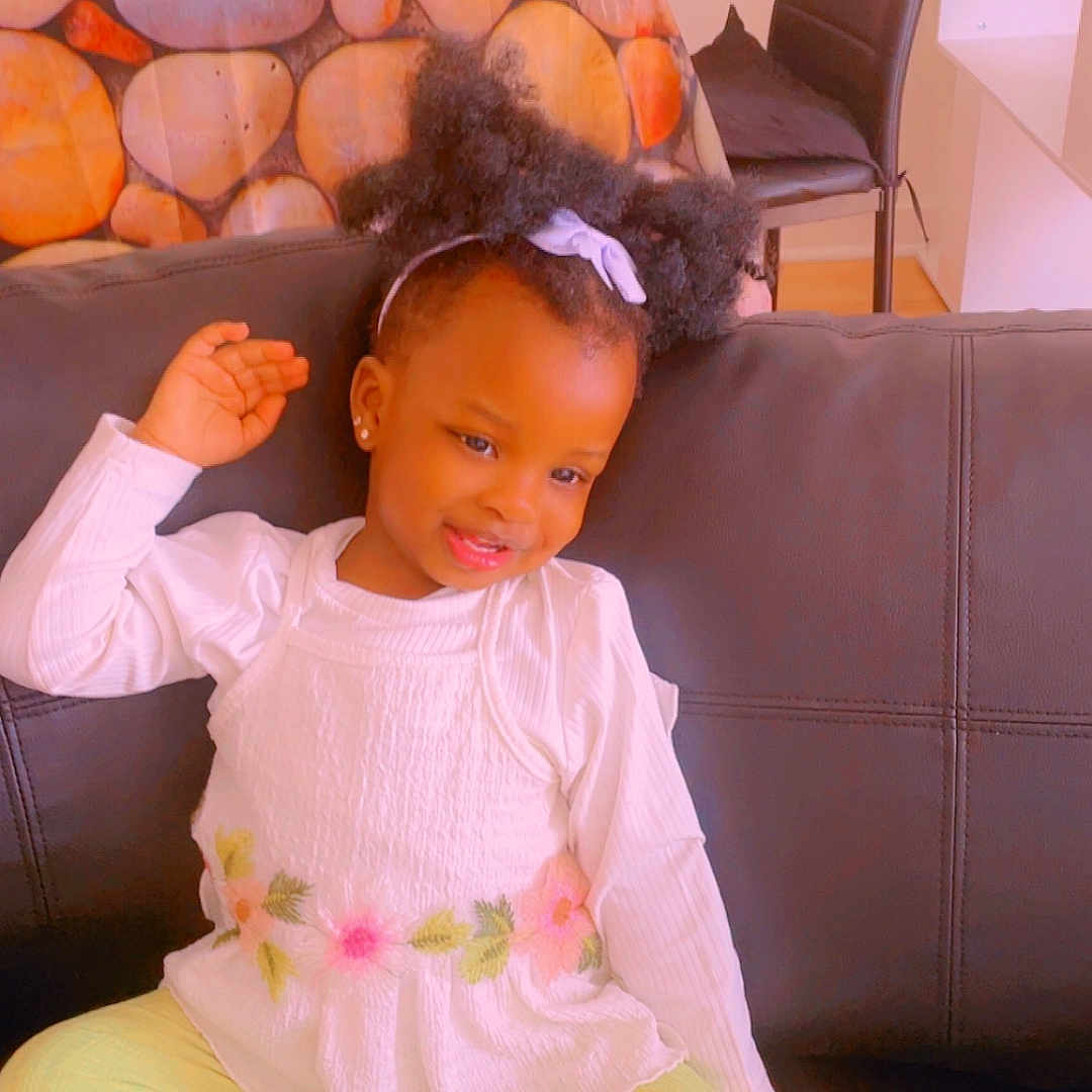 Aita Marieme participe au concours pour gagner de l'argent avec cette photo : toddler, child, sitting, couch, black_couch, white_top, yellow_pants, blue_socks, hair_ribbon, smiling, indoor, table, fruit_basket, bottle, chair, playful, cute, portrait, home, cozy