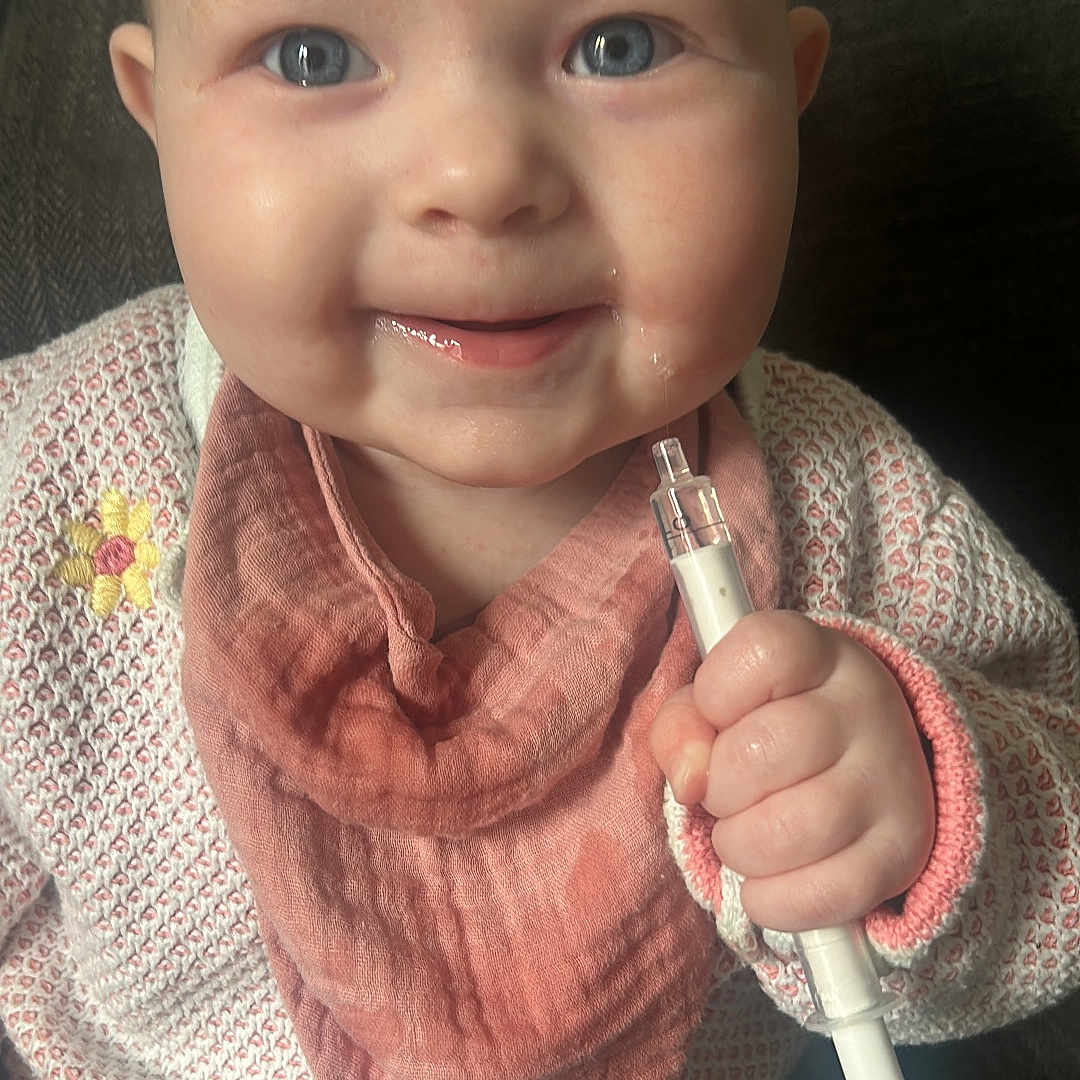 Ivy-Lou is registered to the contest to win money with this photo: baby, blue_eyes, child, closeup, couch, cozy, cute, drool, face, hand, happy, holding_object, indoor, infant, jeans, person, pink_bib, sitting, smiling, sweater