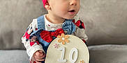 Belle joined the competition — help win amazing prizes! baby, child, couch, cute, denim_overalls, flower, gray, headband, indoors, infant, months, person, portrait, red, sign, sitting, smiling, soft, white_tights, wooden_sign