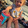 bed, blanket, blue_eyes, child, clothing, comfort, face, flower_pattern, furniture, hair, head, indoor, person, playful, relaxation, sandals, sippy_cup, smile, toddler, toy