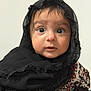 baby, child, face, headscarf, black_clothing, lace, patterned_clothing, wide_eyes, innocent_expression, portrait, person, young_child, cute, curious, indoors, close_up, skin, hand, head, clothing