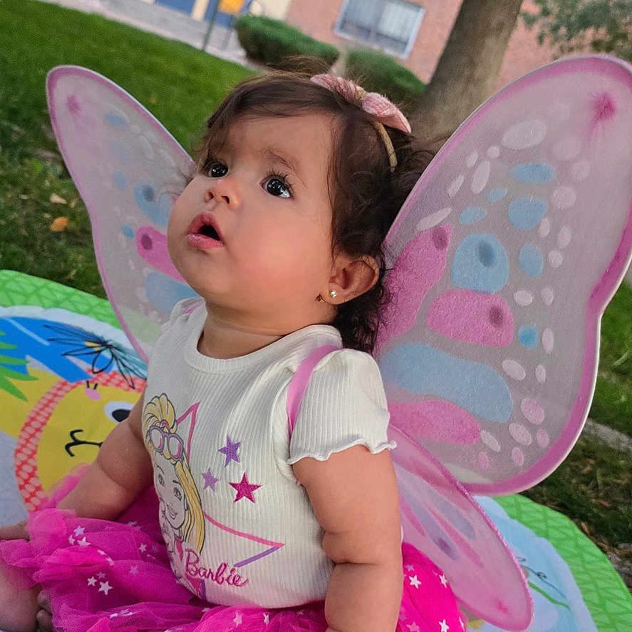 Zoe G.a is registered to the contest to win money with this photo: Barbie, baby, butterfly_wings, child, colorful, curly_hair, daylight, earrings, ears, face, grass, headband, looking_up, outdoor, pink_tutu, play_mat, shirt, sitting, toddler, tree