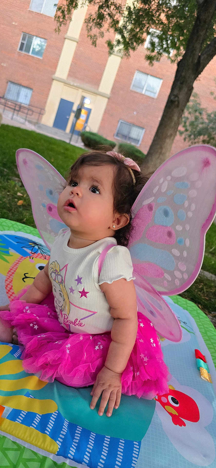 Zoe G.a is registered to the contest to win money with this photo: toddler, child, baby, outdoor, grass, tree, play_mat, pink_tutu, butterfly_wings, headband, curly_hair, face, ears, earrings, shirt, Barbie, colorful, sitting, looking_up, daylight