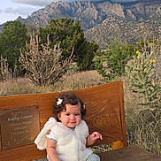 Zoe G.a is registered to the contest to win money with this photo: toddler, child, bench, outdoor, mountains, cloudy_sky, nature, grass, plants, desert, curly_hair, vest, barefoot, portrait, smile, young_child, wood, scenic, daytime, cute