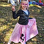child, girl, pink_glasses, black_jacket, pink_skirt, poodle_skirt, playground, outdoor, grass, leaves, smile, peace_sign, shoes, socks, fall_season, play_equipment, happy, posing, daylight, fashion