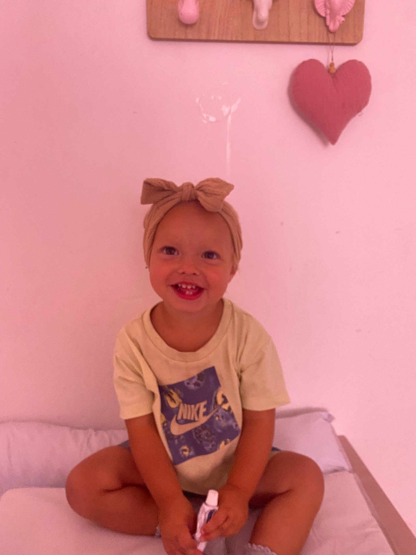 Mya a rejoint le concours — aidez-le/la à gagner de superbes lots ! bed, bow, casual_clothing, child, cute, happy, headband, heart_decoration, holding, indoor, nike, person, pink_wall, portrait, sitting, smiling, toddler, tshirt, tube, young_child