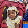 baby, blurred_background, child, crowd, curious, cute, earrings, event, face, hat, indoor, infant, lemon_pattern, person, portrait, public_gathering, red_stroller, seated, stroller, yellow_clothing