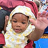 Miracle is registered to the contest to win money with this photo: baby, blue_chair, child, crowd, cute, dress, event, face, happy, hat, indoor, patterned_clothing, person, portrait, red_fabric, seat, smiling, stroller, toddler, waving_hand