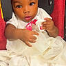 adorable, baby, car_seat, child, closeup, clothing, cute, holding_object, indoors, infant, looking_away, pacifier, person, portrait, red_background, seated, soft_lighting, toddler, white_dress, young_child