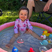 Amelie joined the competition — help win amazing prizes! child, toddler, inflatable_pool, water, toys, rubber_duck, blue_fish_toy, pink_swimsuit, smiling, outdoor, grass, adult, folding_chair, sunlight, summer, playtime, happy, baby, sitting, garden
