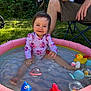 child, toddler, inflatable_pool, water, toys, rubber_duck, blue_fish_toy, pink_swimsuit, smiling, outdoor, grass, adult, folding_chair, sunlight, summer, playtime, happy, baby, sitting, garden