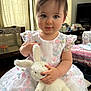 toddler, child, plush_toy, bunny, dress, floral_pattern, indoor, curtain, furniture, playpen, table, television, soft_toy, cute, portrait, baby, person, smile, hand, innocence