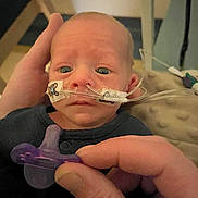 Myles is registered to the contest to win money with this photo: baby, infant, pacifier, hand, medical_tube, nasal_cannula, face, blue_eyes, clothing, person, newborn, indoor, hospital, care, closeup, skin, head, expression, holding, adult_hand