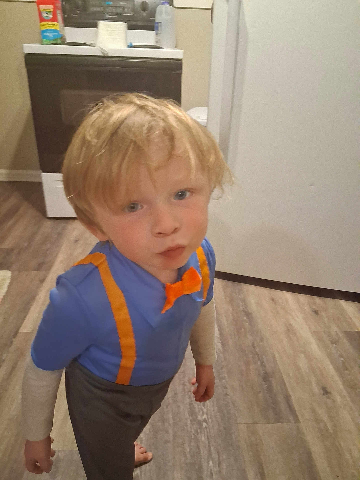 Landin joined the competition — help win amazing prizes! child, boy, blonde_hair, blue_shirt, orange_suspender, orange_bow_tie, barefoot, kitchen, floor, wood_floor, stove, refrigerator, curious_expression, standing, indoor, casual_clothing, pout, young, person, home