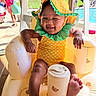 baby, infant, child, smiling, yellow_hat, pineapple_pattern, swimsuit, inflatable_chair, teddy_bear_print, poolside, outdoor, sunny, feet, happy, summer, water, people, background, hat, playful