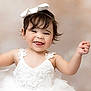 baby, toddler, girl, smile, portrait, white_dress, bow_headband, lace, tulle, cute, adorable, dark_hair, teeth, ears, earrings, raised_hand, studio_background, soft_lighting, happy, person
