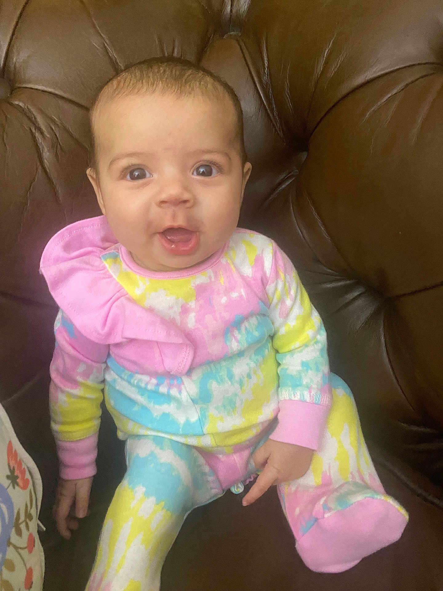 Renesme is registered to the contest to win money with this photo: baby, child, infant, tie_dye, colorful_clothing, sitting, couch, leather, brown, cute, wide_eyes, expression, indoors, portrait, person, happy, small_hands, comfortable, young, cozy