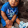 Leonidas is registered to the contest to win money with this photo: baby, child, portrait, sitting, couch, indoors, blue_outfit, curly_hair, smiling, serious_expression, sneakers, white_shoes, cartoon_shirt, fashion, living_room, furniture, window, blinds, natural_light, close_up